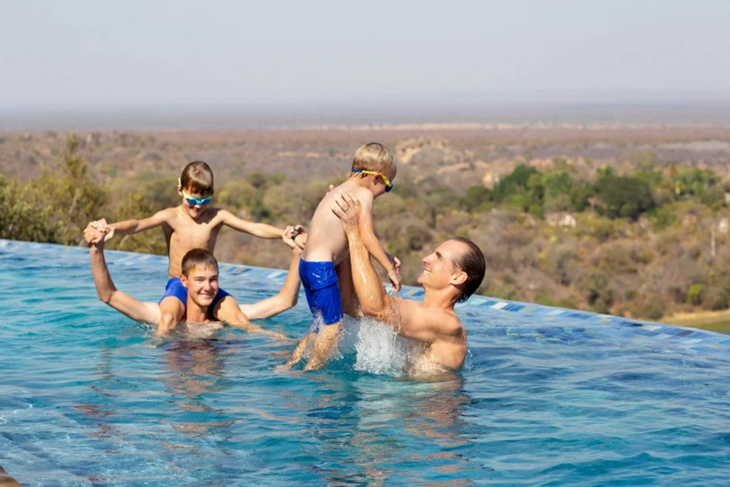 A man and three children playing together in an infinity pool overlooking a vast natural landscape.