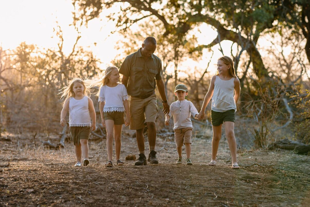 A safari guide walking hand-in-hand with four young children along a dirt path in the bush at sunset.
