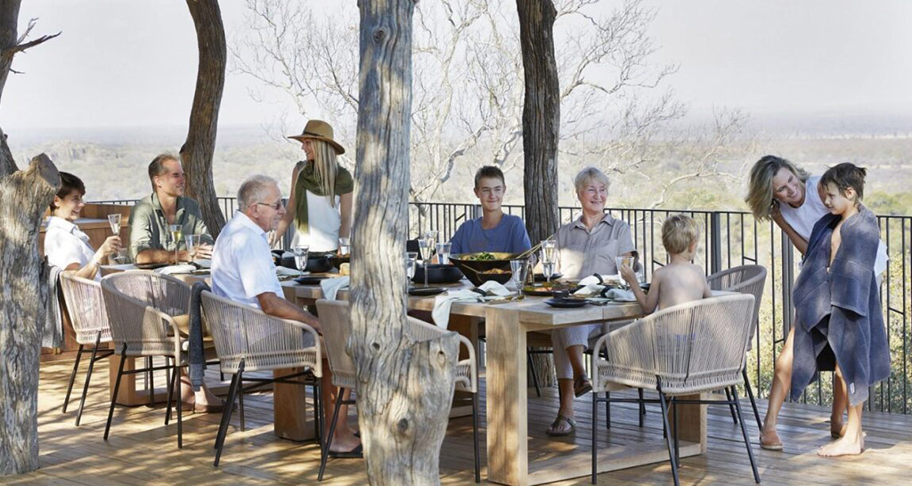 A multigenerational family enjoying a meal together on a wooden deck overlooking a scenic wilderness view.