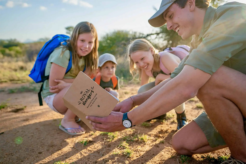 A safari guide showing a field guide book labelled "Royal Rangers" to three smiling children outdoors.
