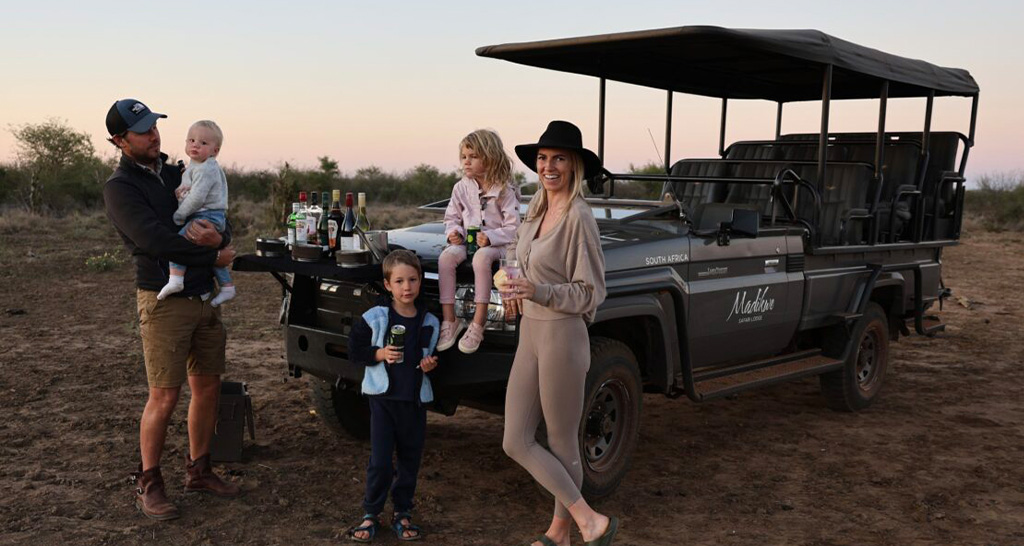 A family with three children stands beside a safari vehicle in South Africa during sunset, with a drink station set up on the front of the vehicle.