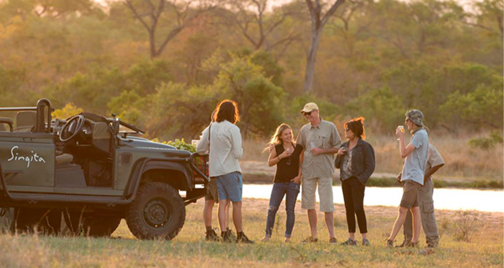 A group of people standing beside a safari vehicle labelled "Singita," enjoying drinks at sunset in a natural setting.