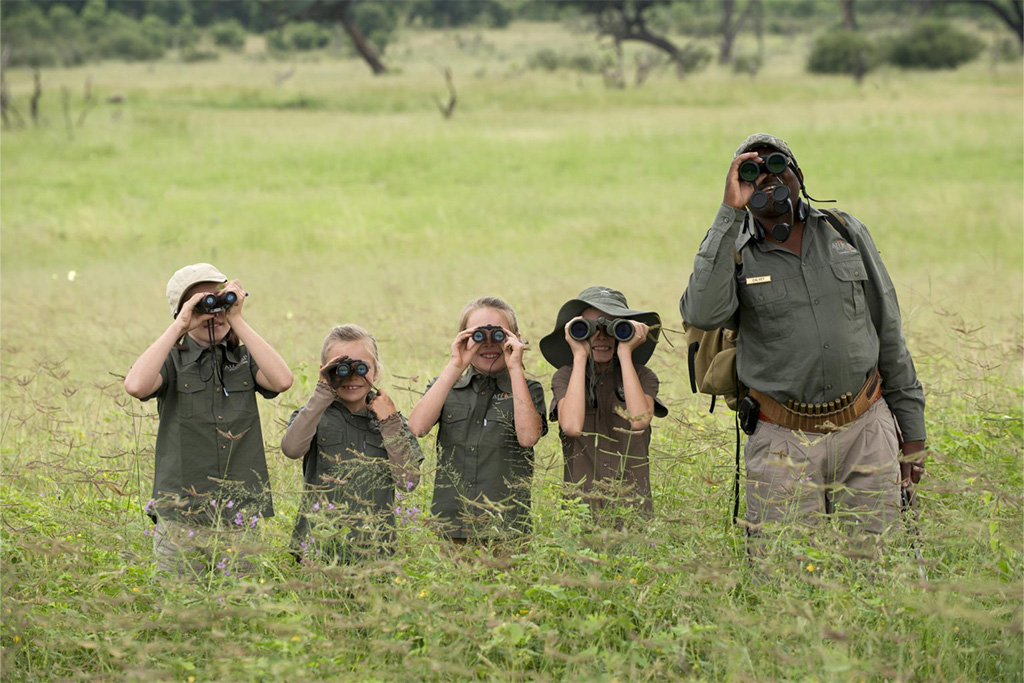 A group of four children and a safari guide standing in tall grass, all looking through binoculars in the same direction.