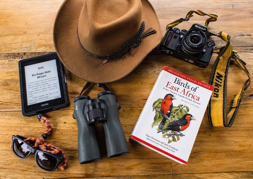 Safari packing essentials displayed on a table including a sun hate, binoculars, camera, sunglasses, and birding guide.