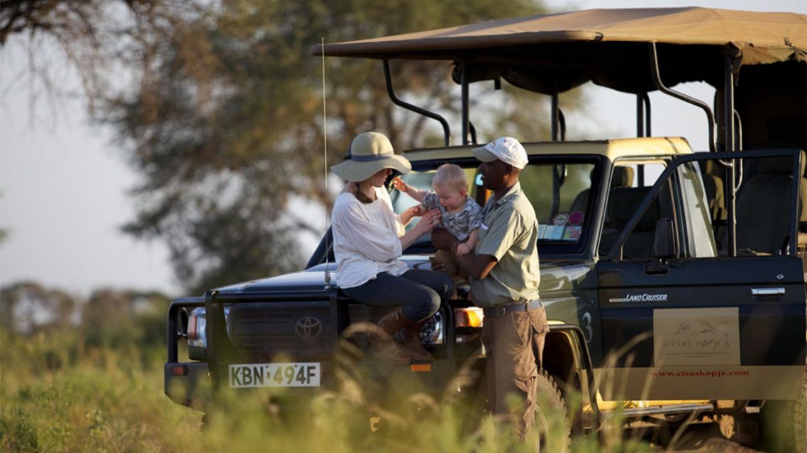 A safari guide hands a baby to a woman sitting on the hood of a safari vehicle, with open grasslands and trees in the background.