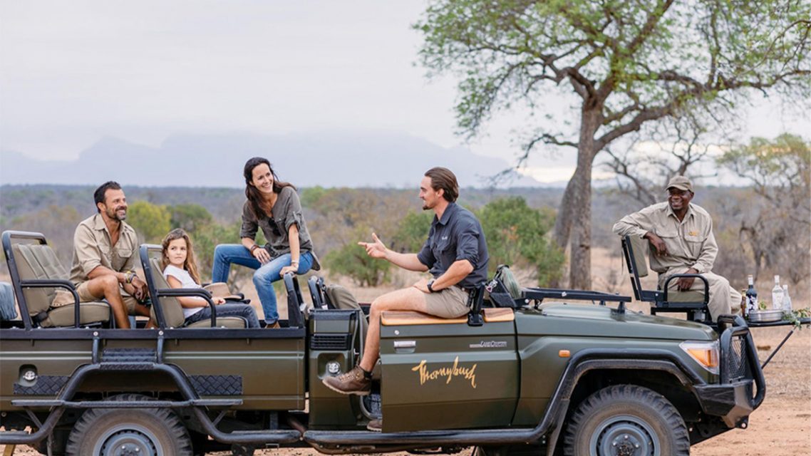 A family sits in an open safari vehicle talking with guides during a break in the African bush, with drinks set up on a small table at the rear of the vehicle.